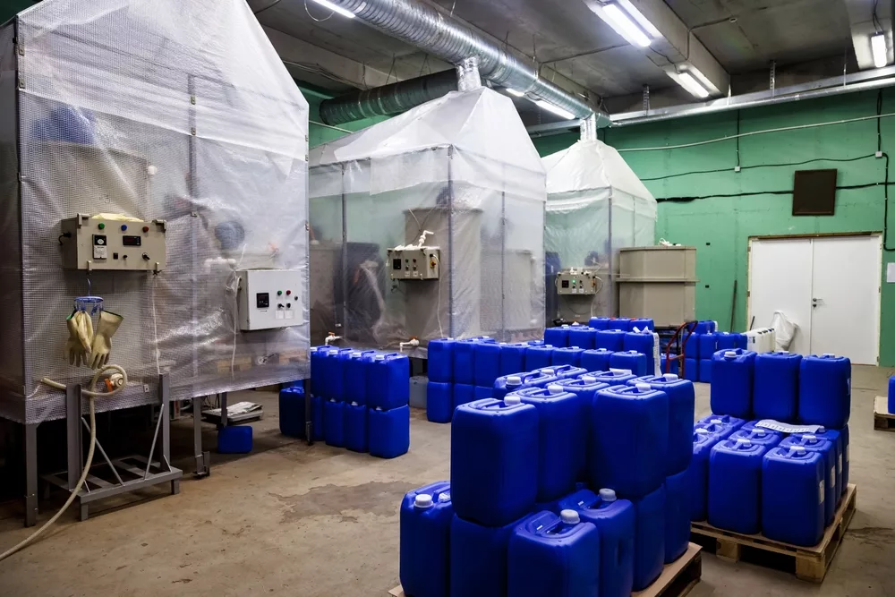 Industrial room with plastic-covered machinery and large air ducts, surrounded by numerous blue chemical barrels arranged in stacks, conveying a functional, utility-focused atmosphere.