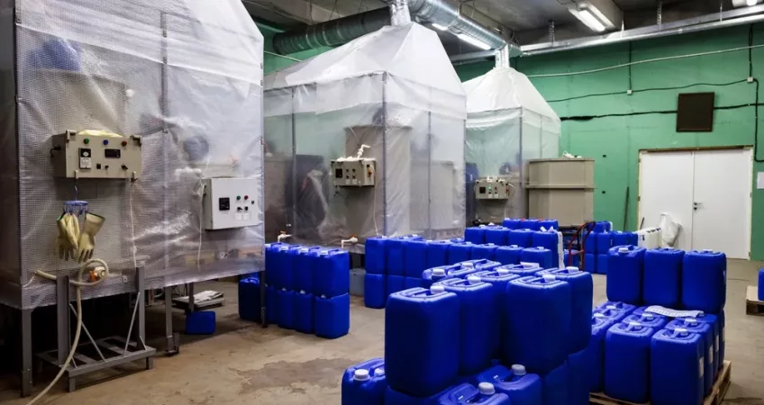 Industrial room with plastic-covered machinery and large air ducts, surrounded by numerous blue chemical barrels arranged in stacks, conveying a functional, utility-focused atmosphere.